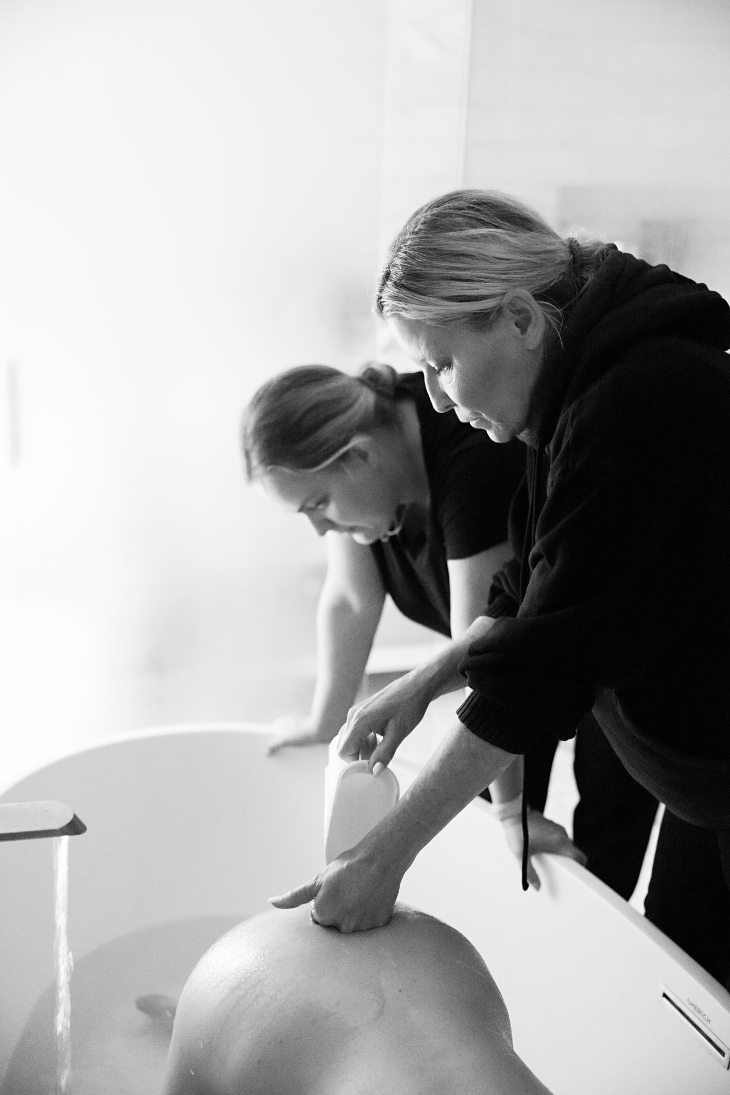 Midwife pouring water over pregnant womans back leaning over white bathtub.