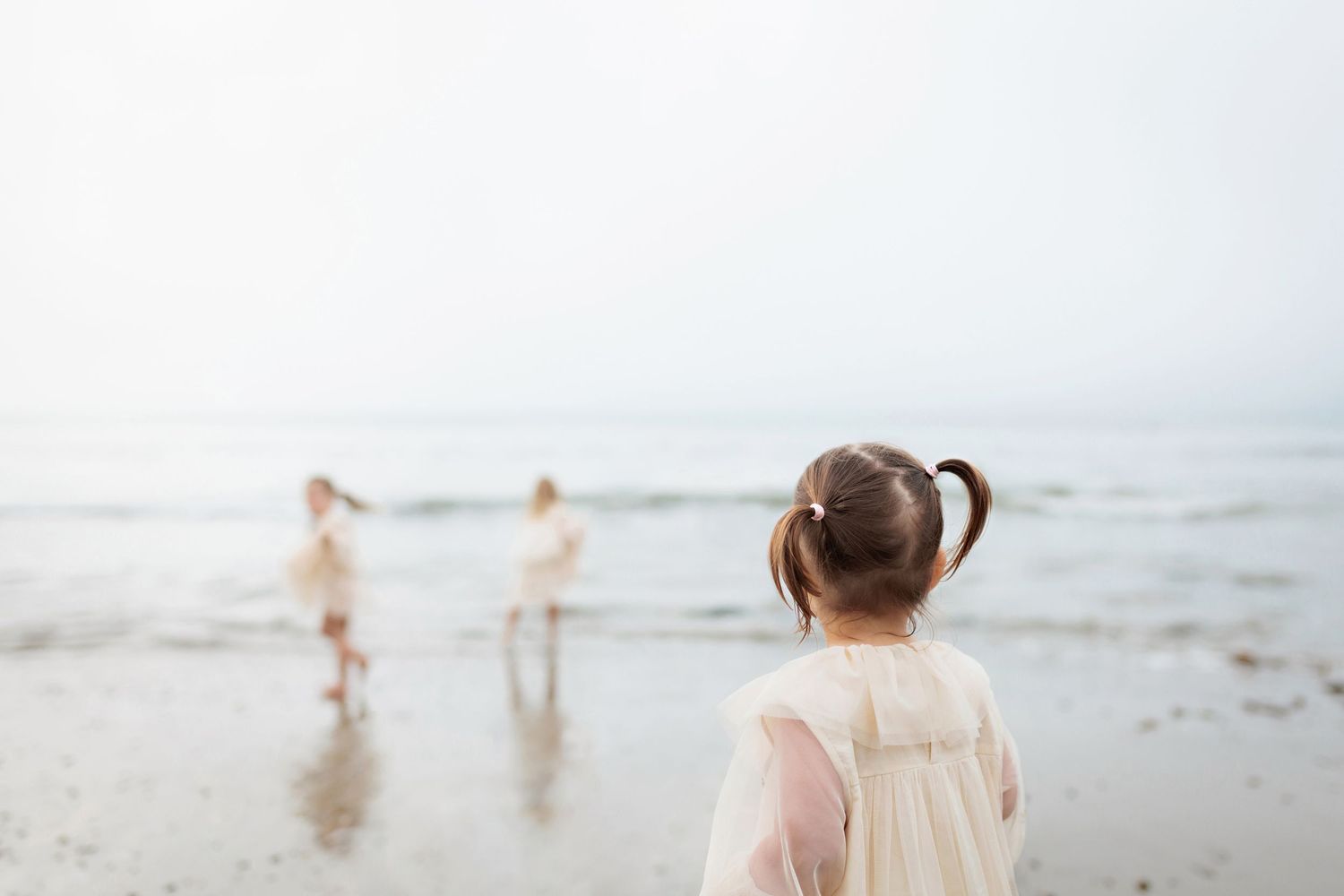 Young girl with pigtails in white dress watching two children play at the ocean shoreline.
