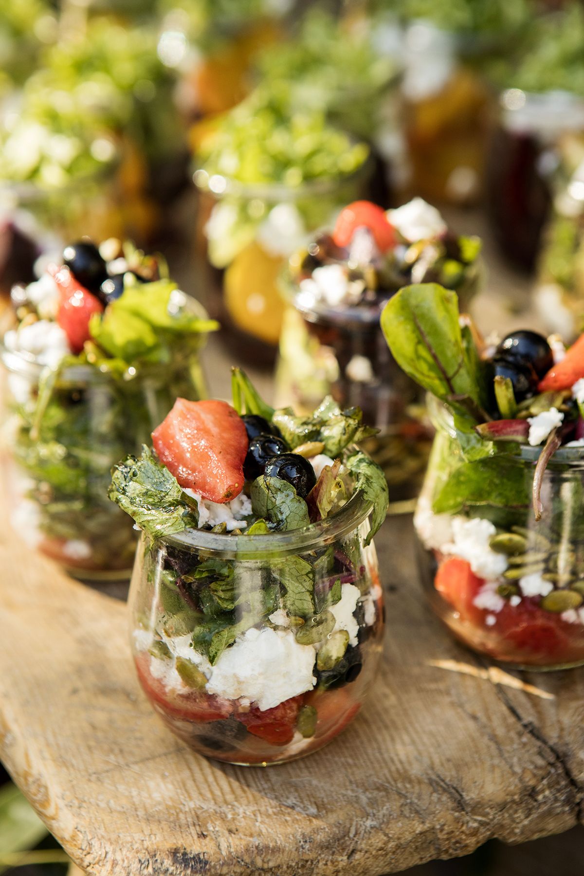 Individual glass cups filled with layered Greek salad featuring watermelon, feta, olives, and greens on a rustic wood surface.