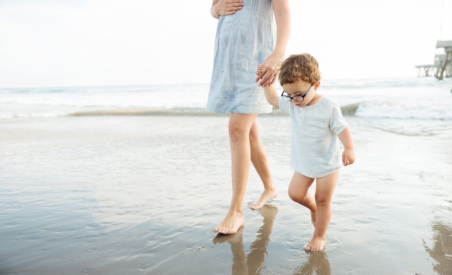 A toddler walks barefoot on a sandy beach beside a pregnant woman in a light blue dress near the ocean.