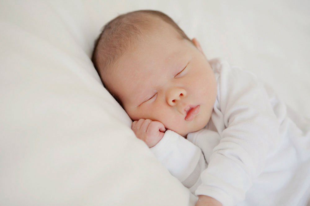 Peaceful newborn baby sleeping on white bedding, wearing a white onesie with tiny hands tucked under cheek.