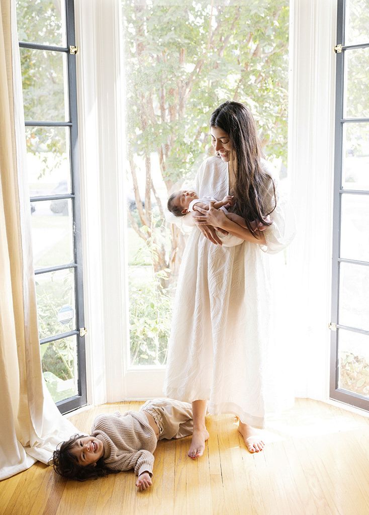 Mother in white dress holds newborn by French doors while toddler plays on hardwood floor in bright airy home.