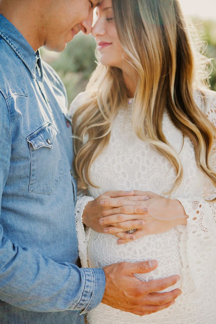 Couple tenderly embracing during maternity photo shoot, hands cradling baby bump, wearing denim and white lace dress.