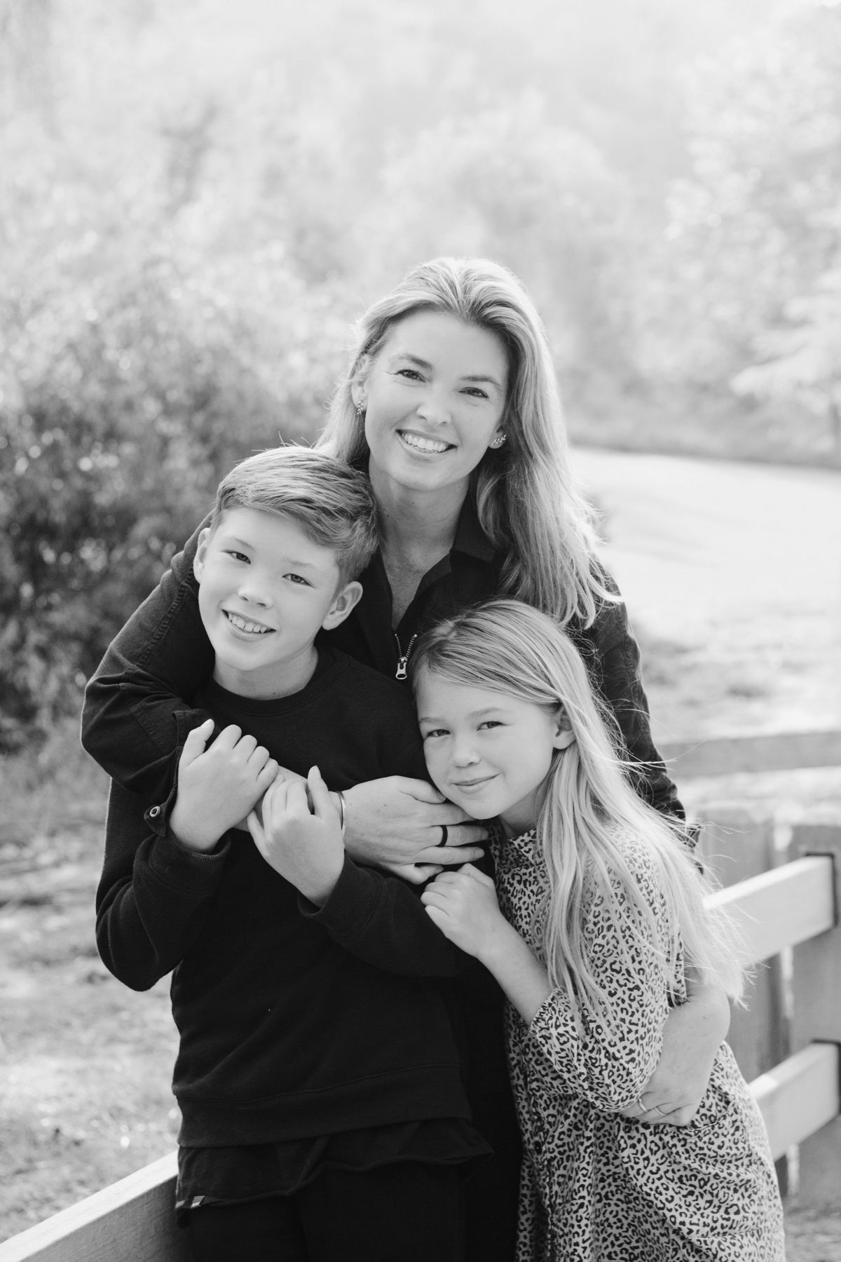 Black and white portrait of a smiling mother hugging her two children outdoors near a wooden fence.