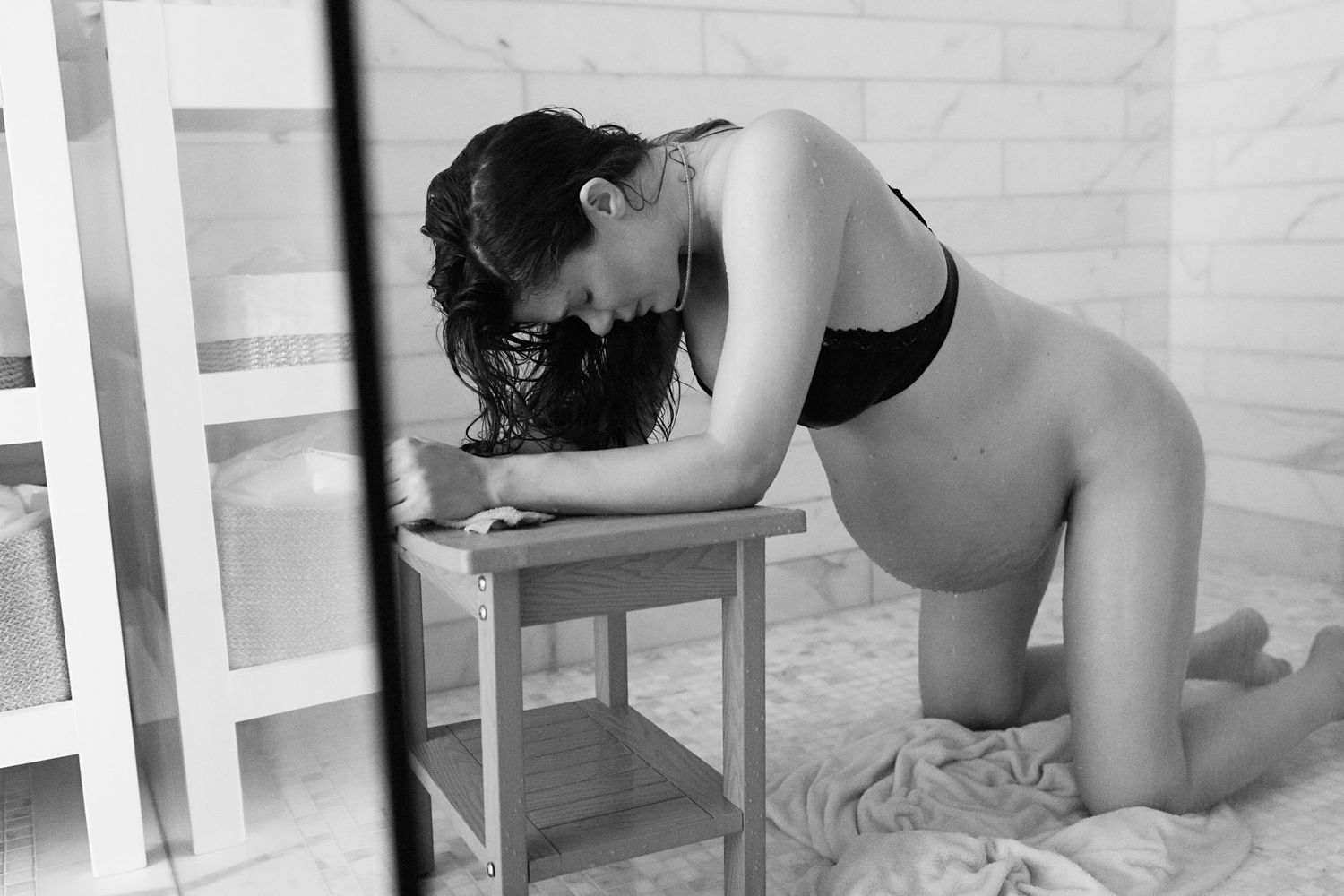 Pregnant woman in black bikini kneeling beside wooden stool in bathroom.