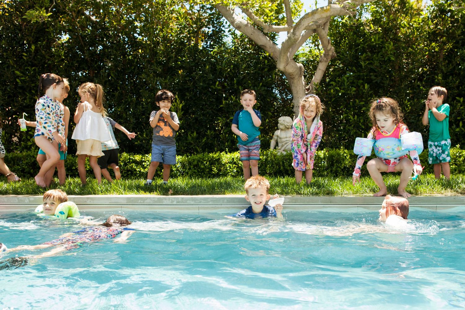 Children enjoying a sunny backyard pool party, with kids swimming and standing poolside in colorful summer outfits.