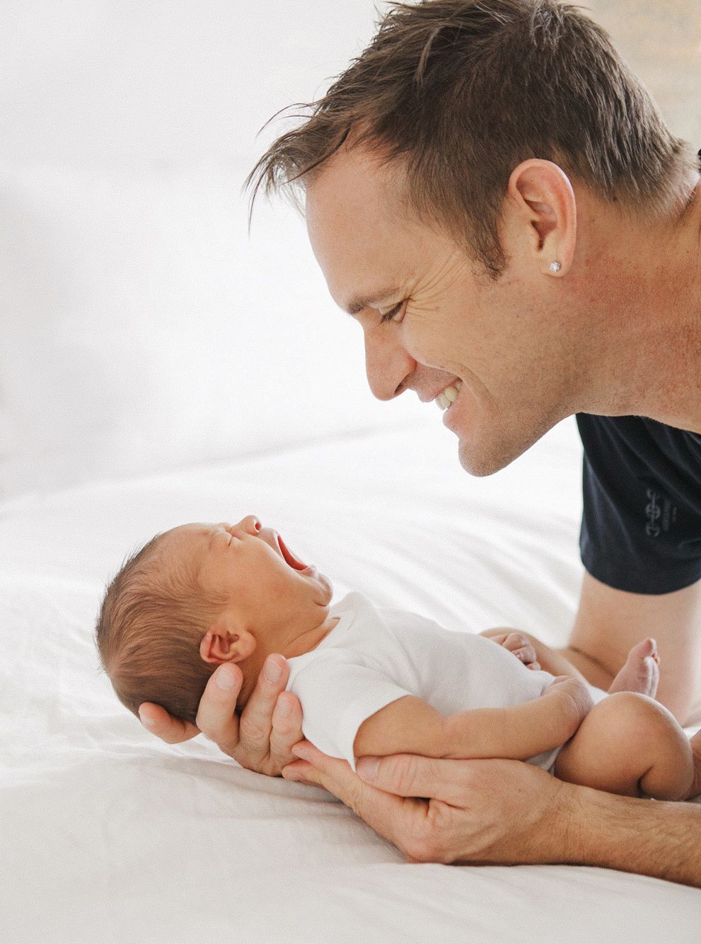 Smiling father holding his newborn baby on a white bed, sharing a joyful bonding moment.