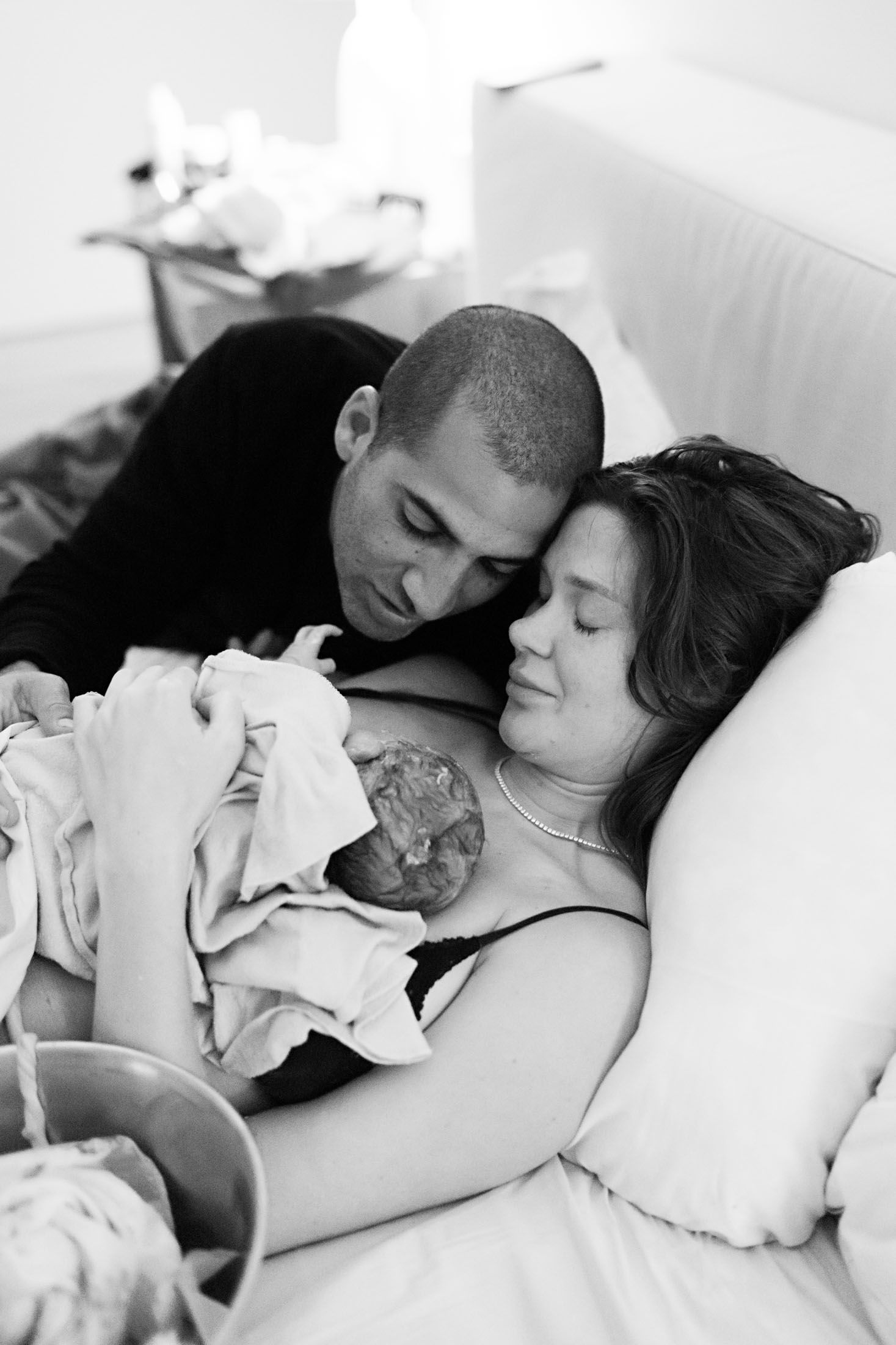 Father leans close to embrace mother holding newborn baby after home birth delivery.