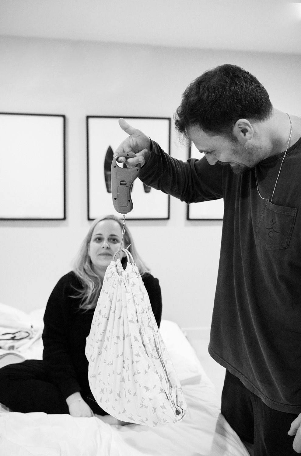 Man holds up baby bag while blonde woman sits on hospital bed, black and white newborn photo.