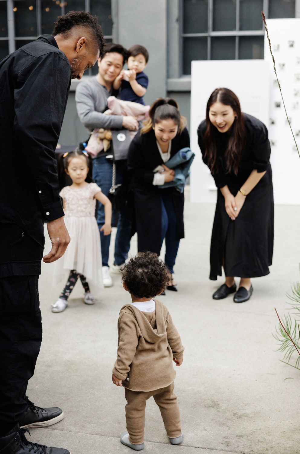 A toddler in a tan hoodie stands before smiling family members during a Korean dol birthday celebration outdoors.