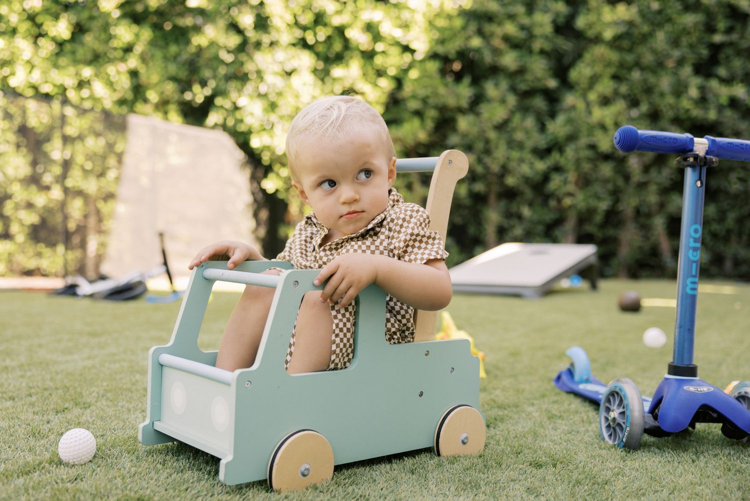 Toddler boy playing with a wooden toy push cart on green grass in a backyard with a blue scooter nearby.