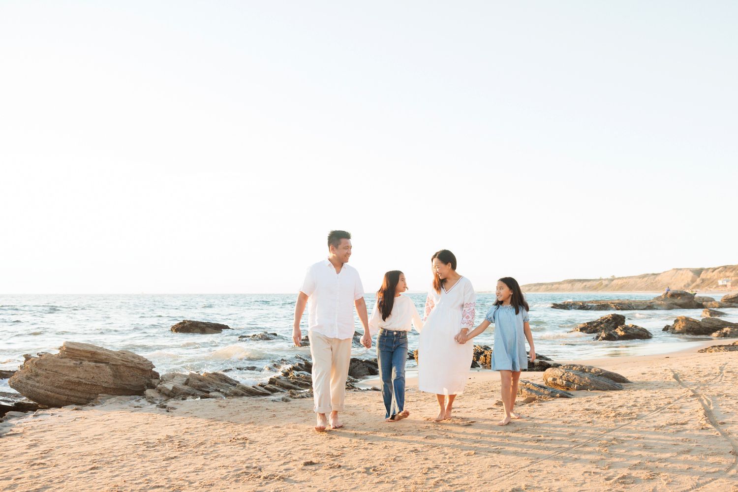 Family of four walking hand-in-hand on a sunny beach with rocky shores and calm ocean waves in the background.