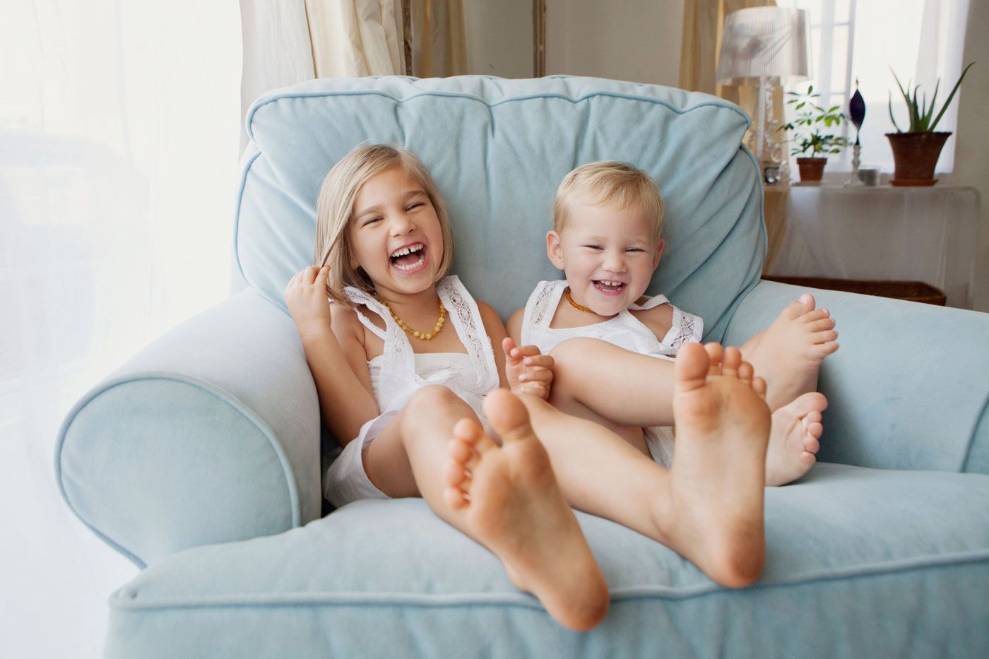 Two laughing children sitting barefoot on a light blue armchair in a bright living room.