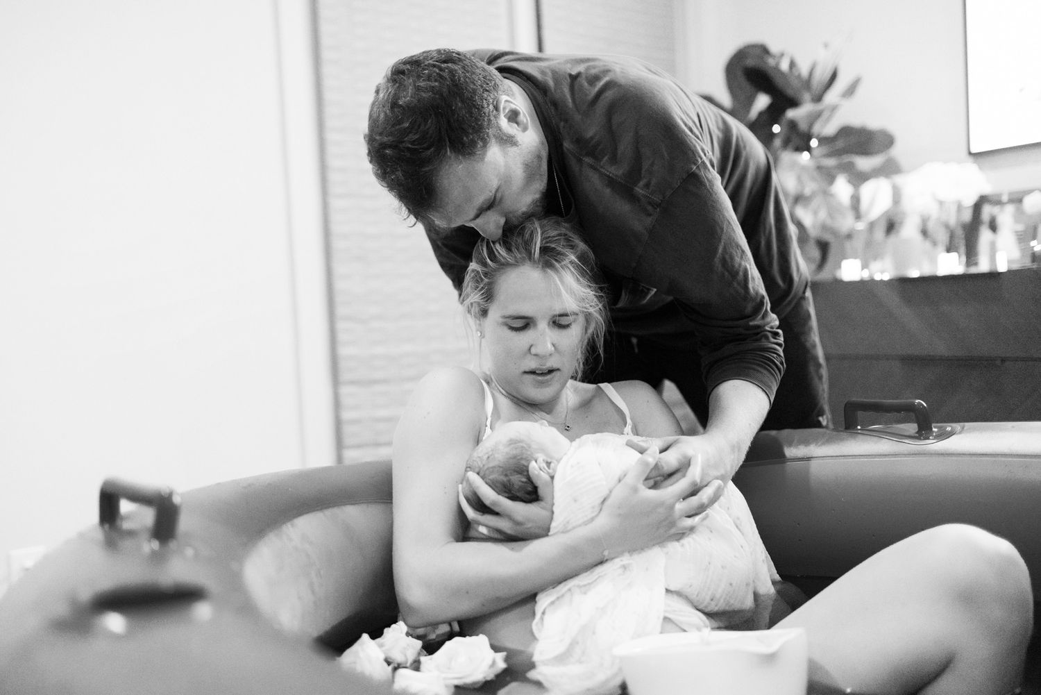 Black and white image of a father embracing mother and newborn baby beside a birth pool after water birth.