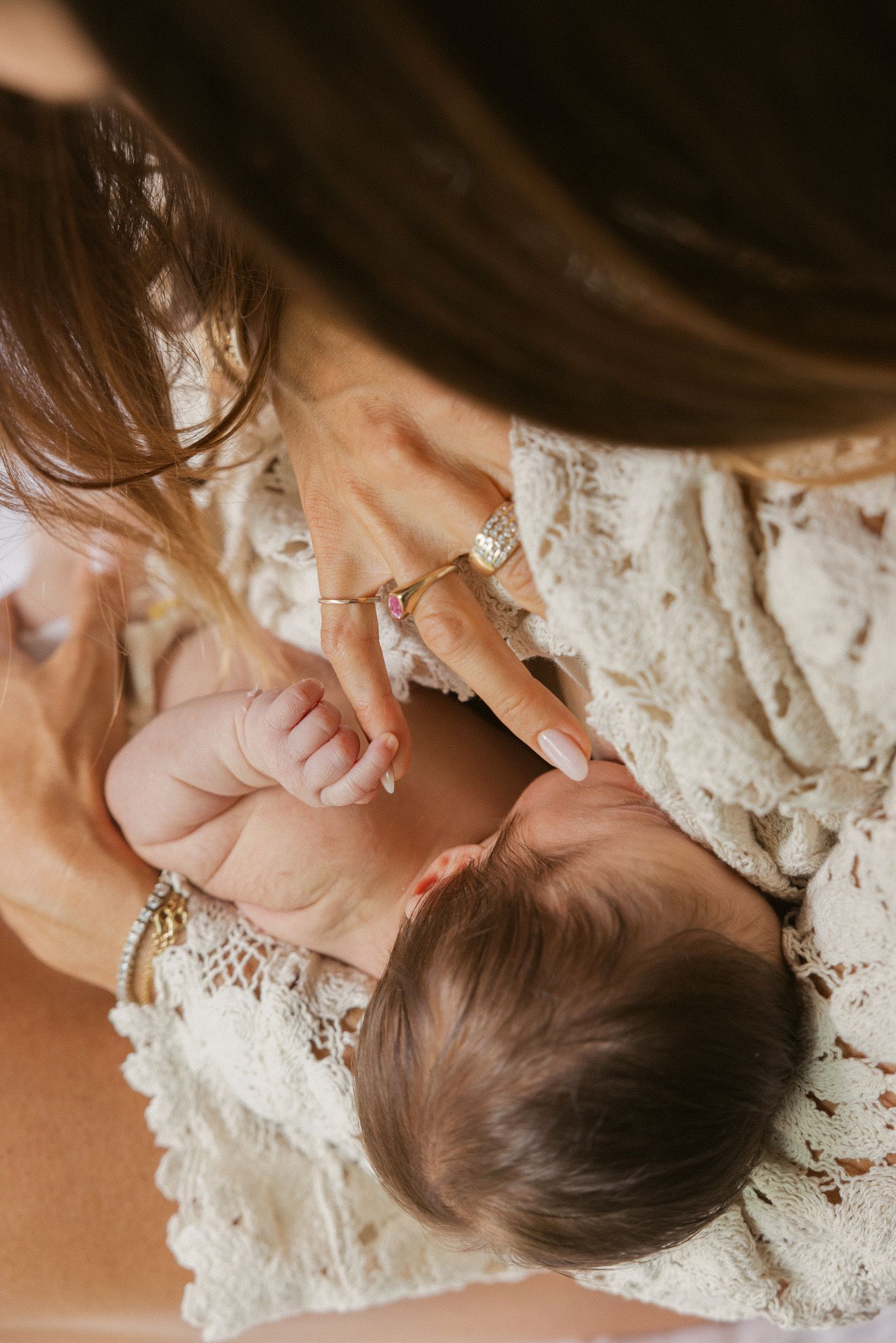 Mother holding newborn baby while wearing gold rings and bracelets, lace dress, tender bonding moment.