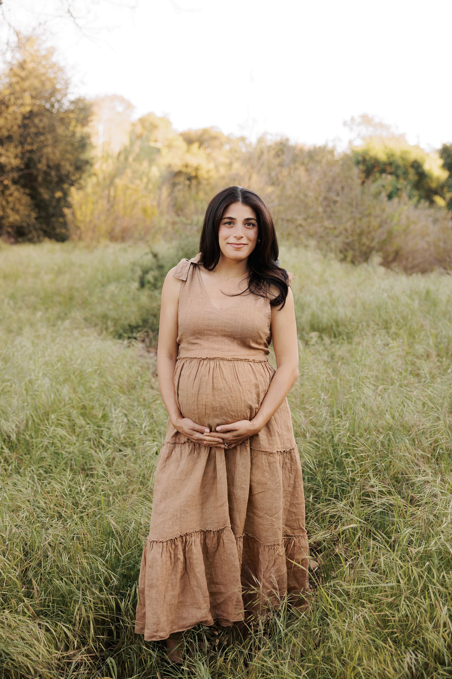 Pregnant woman in tan linen dress standing in green field, cradling baby bump, outdoor maternity photo.