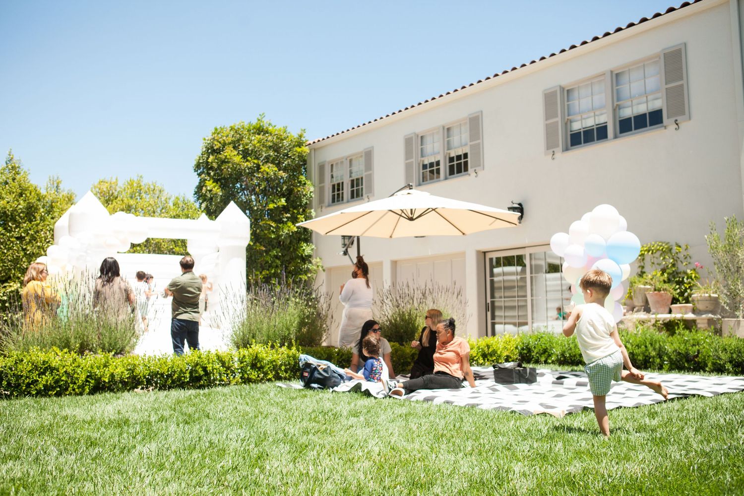 Children playing on a sunny lawn with a white bouncy castle and umbrella during an outdoor birthday party.