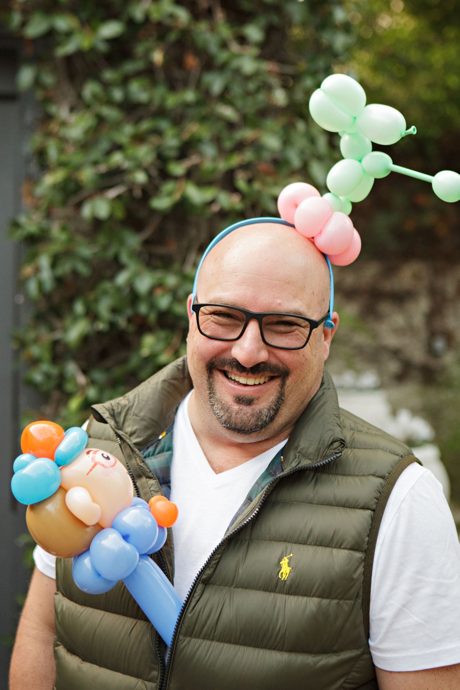 Smiling bald man wearing glasses with colorful balloon animals on his head and shoulder at an outdoor celebration.