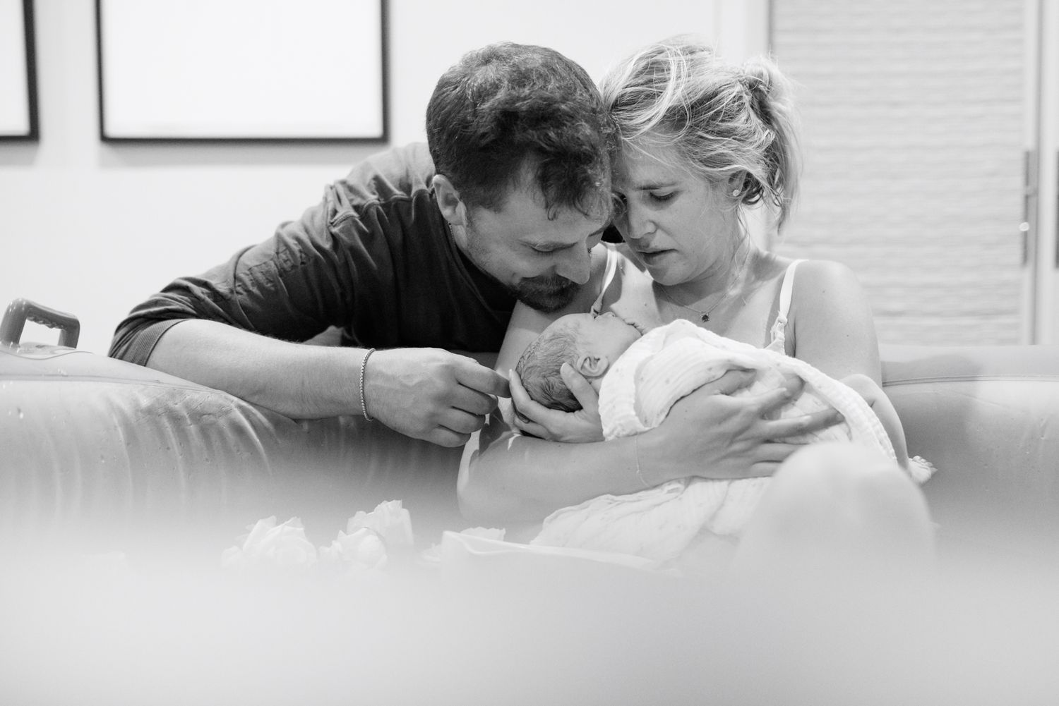 Black and white close-up of new parents lovingly gazing at their swaddled newborn baby together on a couch.