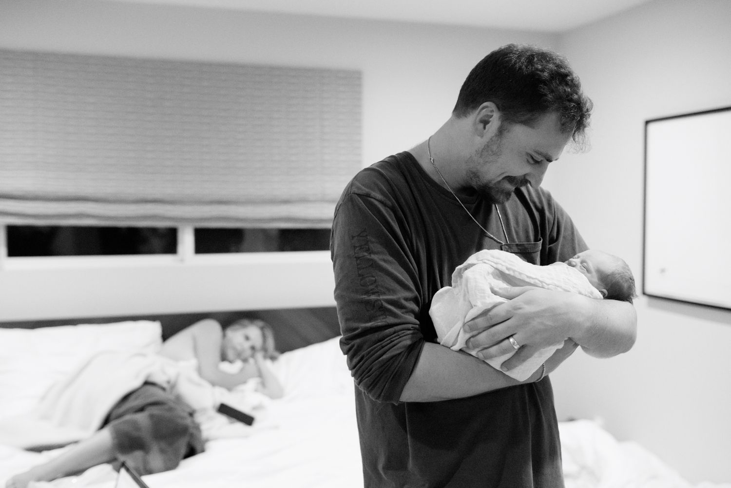 Father holds sleeping newborn baby while mother rests on bed in background, black and white photo.