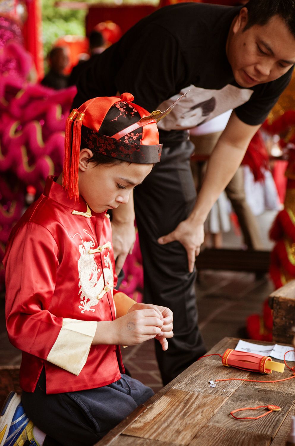 A man helps a child dressed in traditional red Chinese New Year attire with a dragon emblem at a festive celebration.