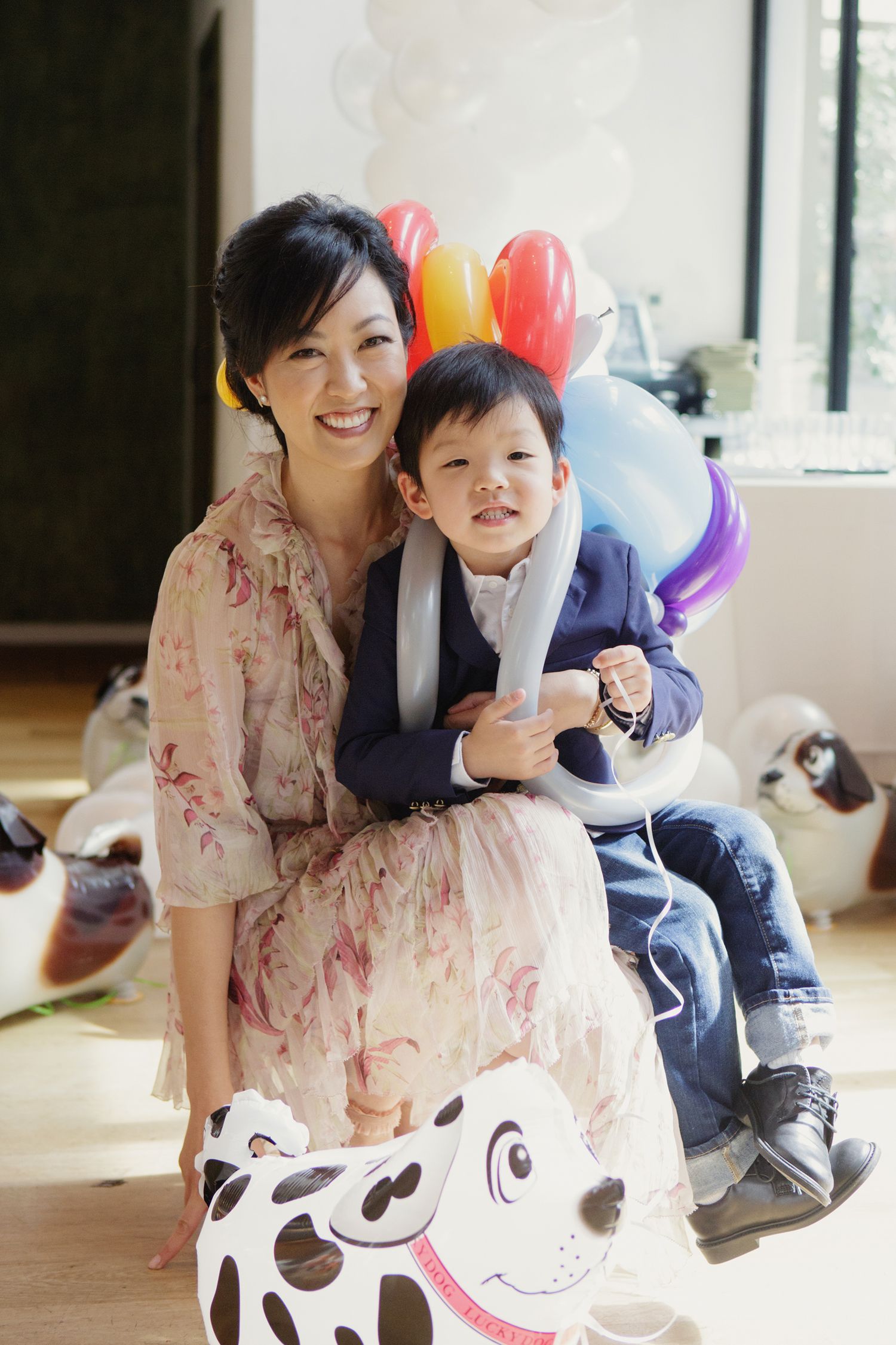 Smiling Asian mother poses with young son on toy dalmatian dog surrounded by colorful balloons.
