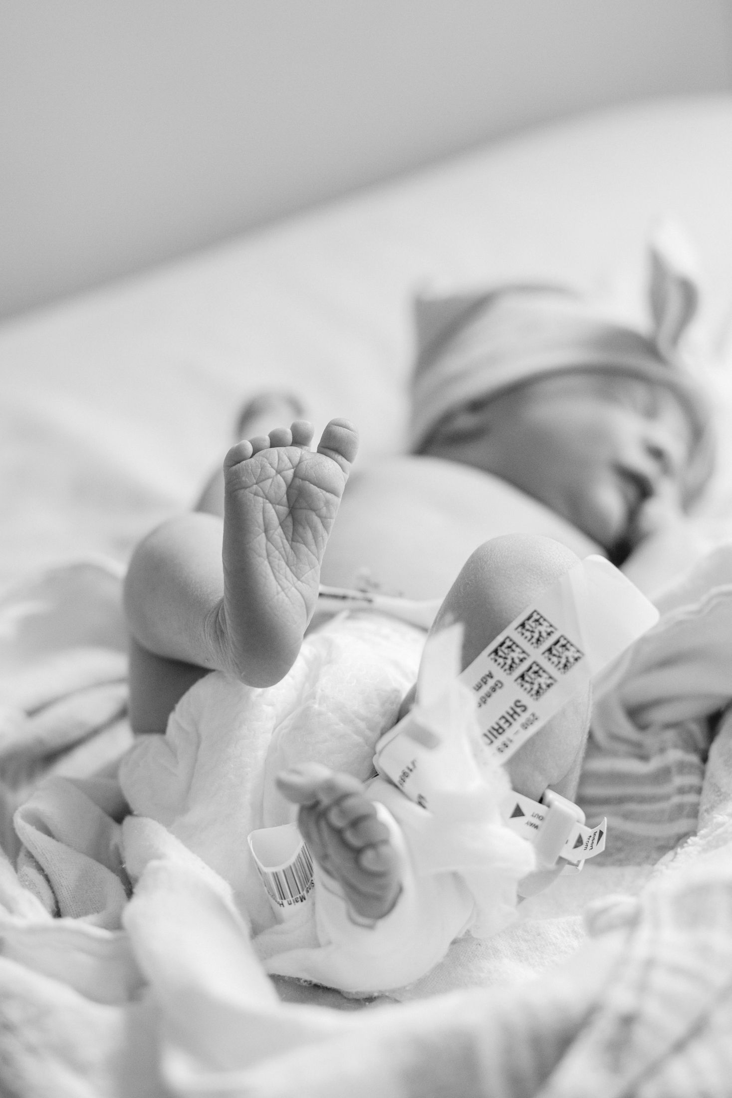 Black and white photo of a sleeping newborn baby in a diaper with hospital ID bands on ankle in a hospital bed.