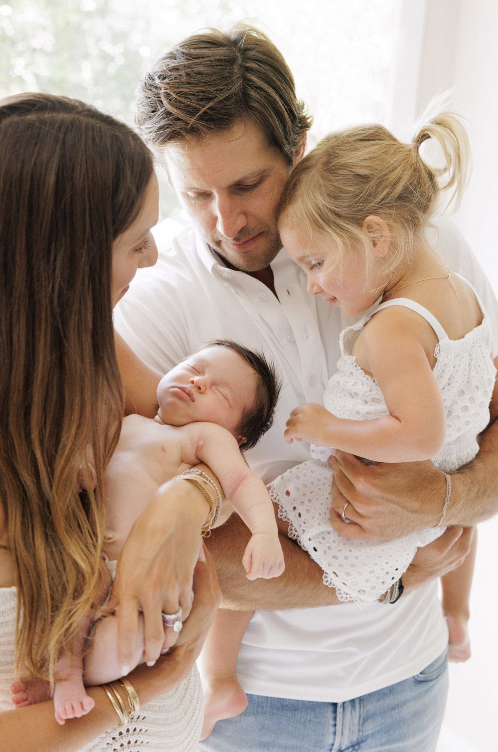 Family of four with parents holding newborn baby and toddler girl, sharing a tender moment together.