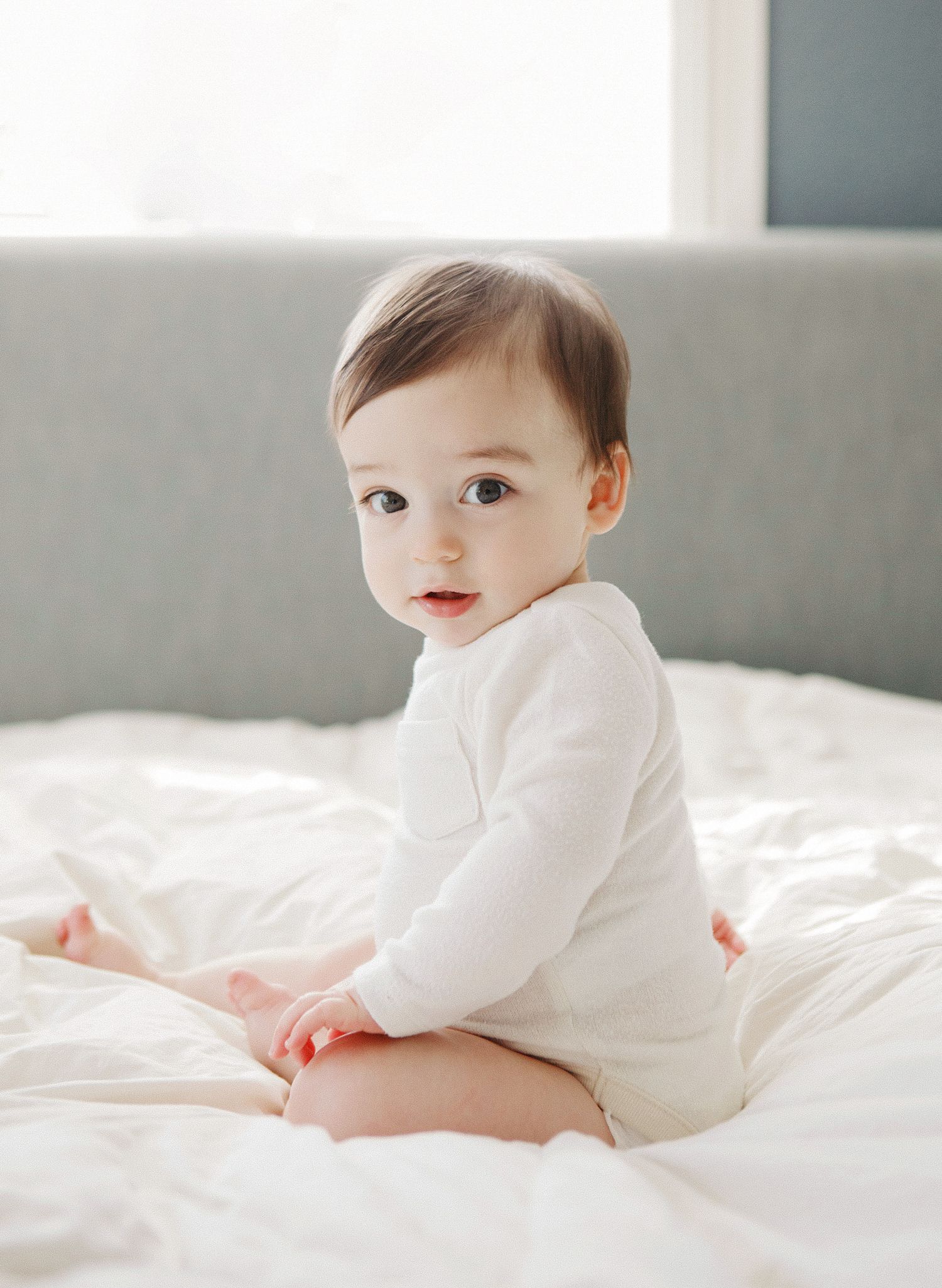 Adorable baby in white onesie sitting on a bed with white bedding, looking back over shoulder.