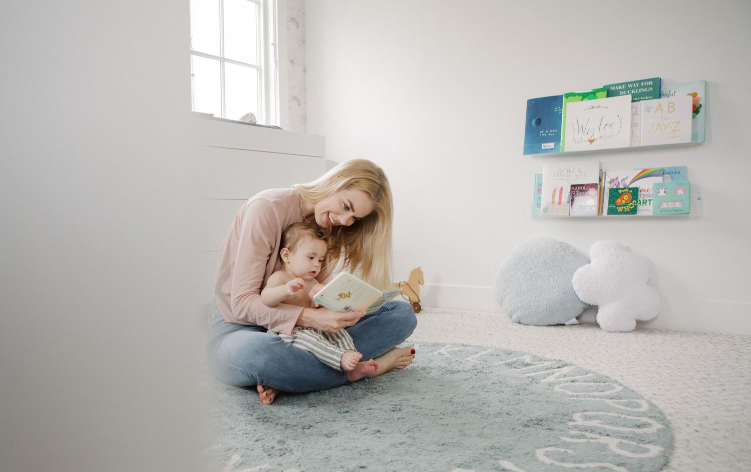 Mother reading a picture book with her toddler on a soft rug in a bright, minimalist nursery room.