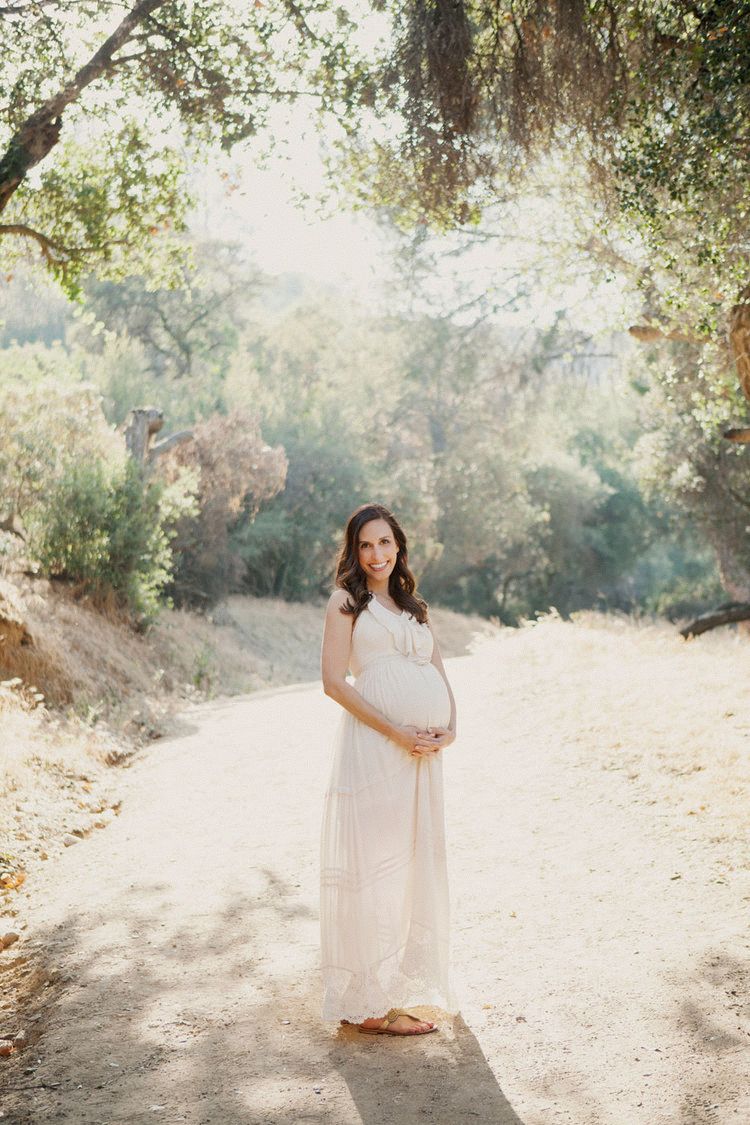 Pregnant woman in white maxi dress cradling baby bump on sunny dirt path surrounded by lush green trees.
