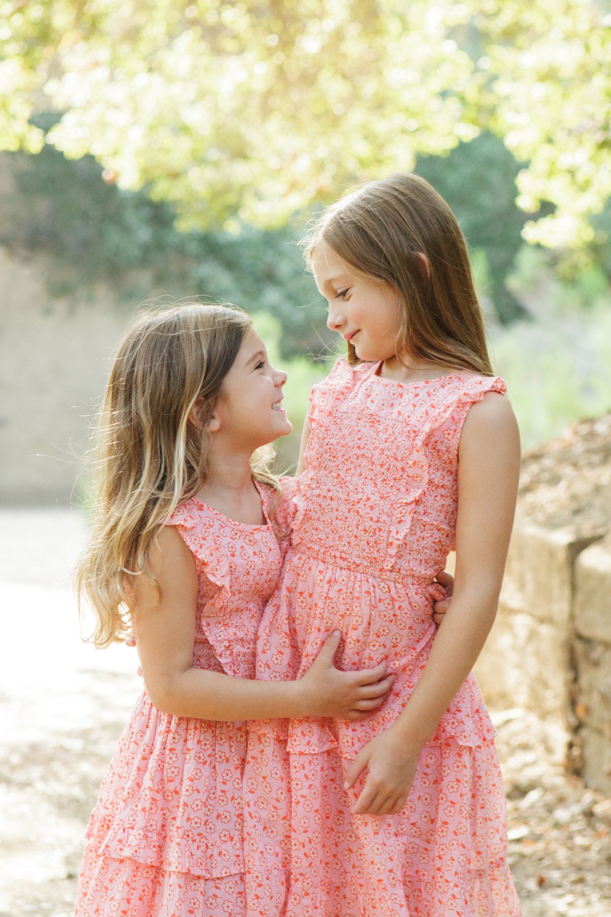 Two young girls in matching pink floral dresses smile at each other outdoors in a sunlit garden setting.