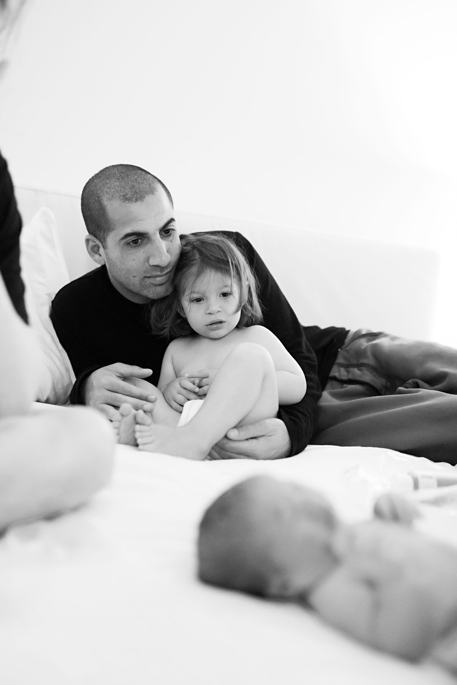 A father tenderly holds his toddler while gazing at a newborn baby on a white bed in black and white.