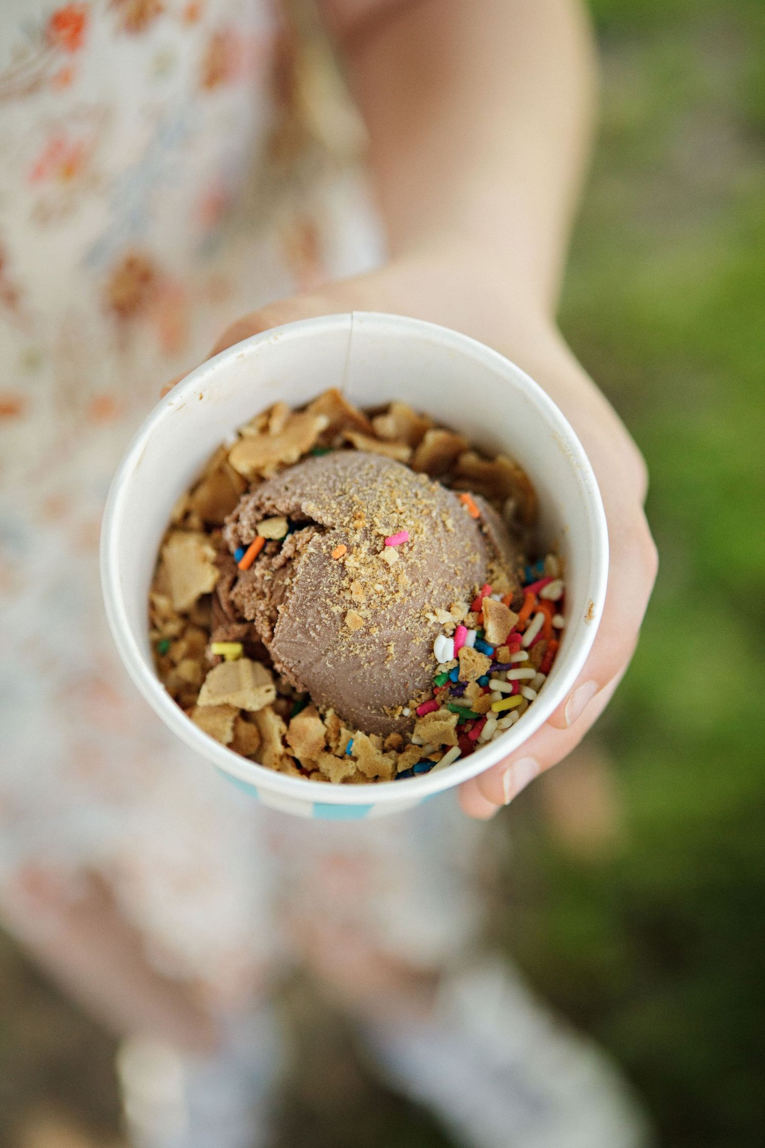 Child holding a cup of chocolate ice cream topped with colorful sprinkles and cookie crumbles outdoors.