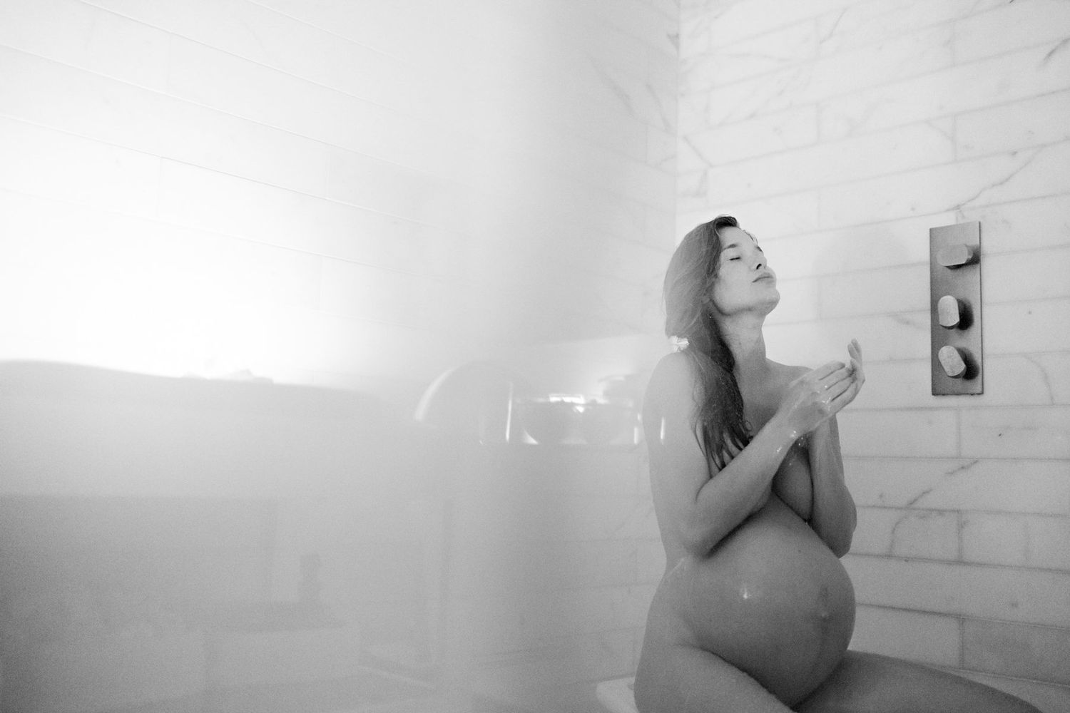 Black and white photo of a pregnant woman kneeling in a steamy shower, hands clasped, eyes closed peacefully.