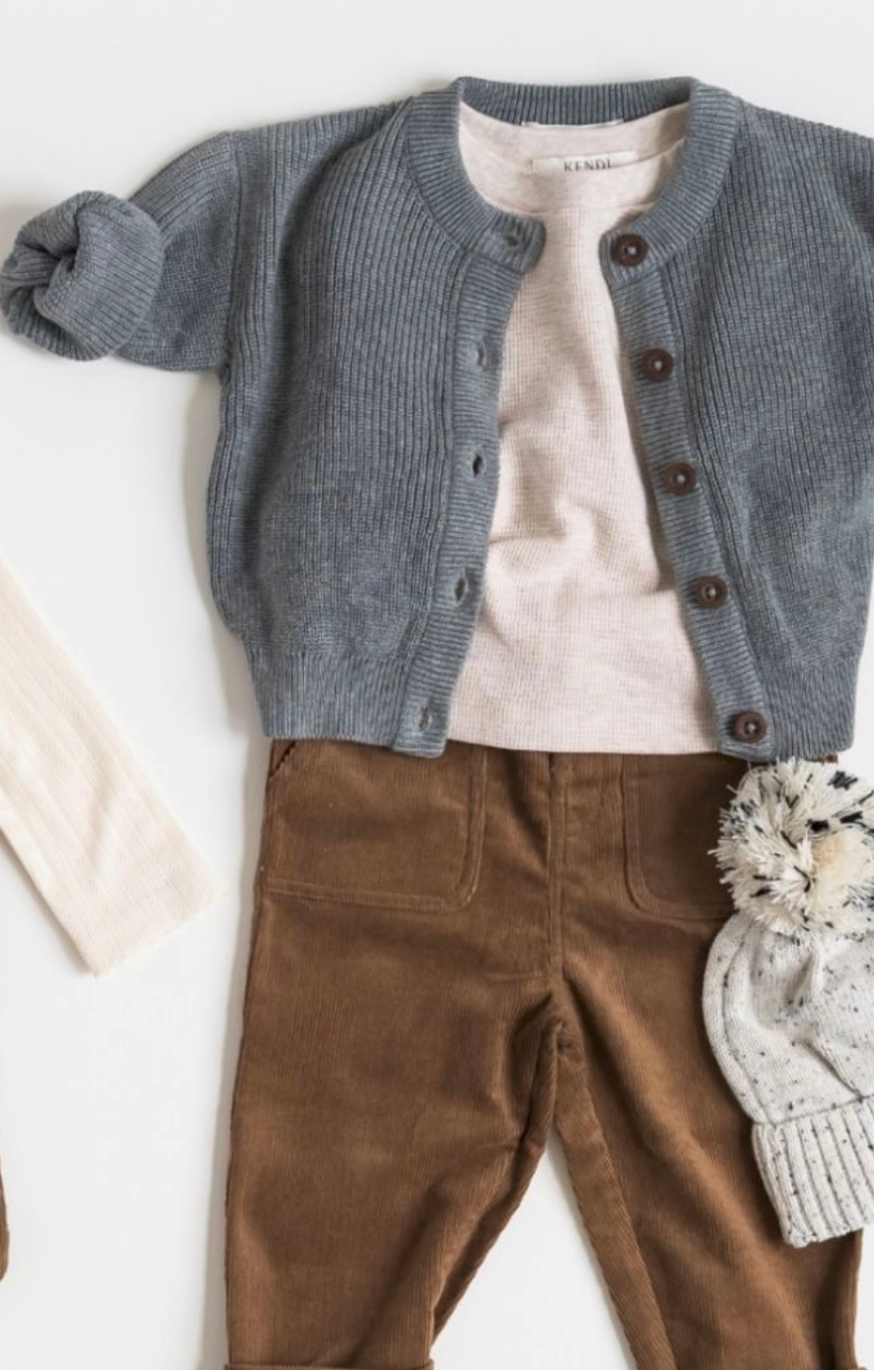Baby outfit flat lay with gray knit cardigan, beige tee, brown corduroy pants, cream tights, and pom-pom hat.