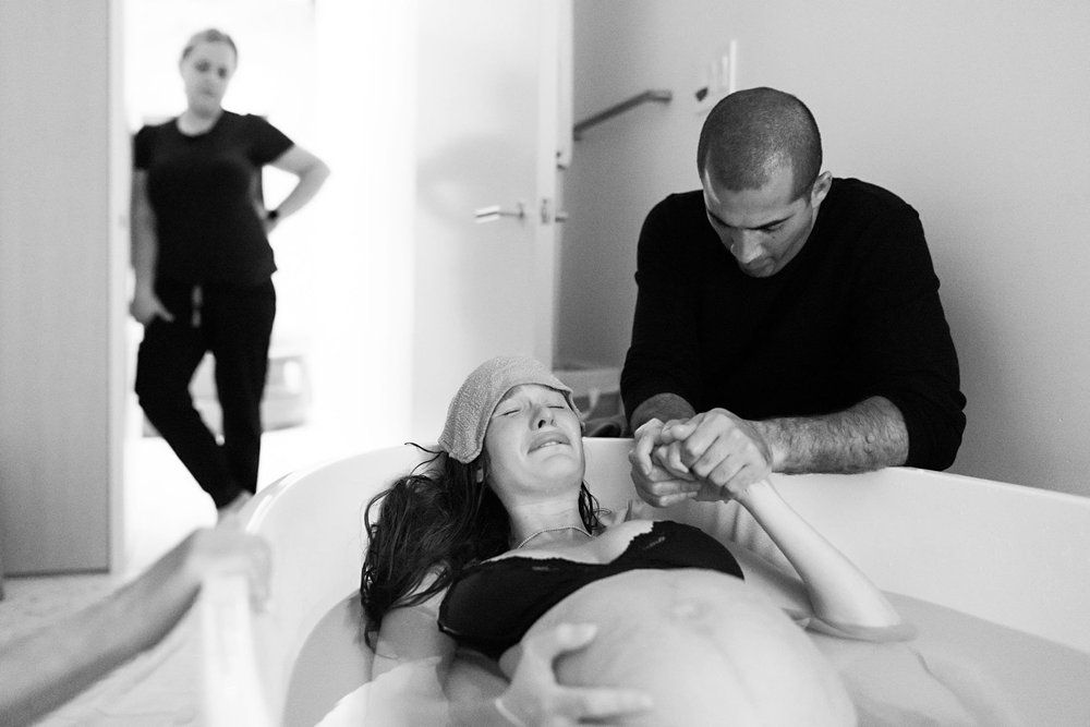 Man comforting woman during water birth labor, midwife in background, black and white.