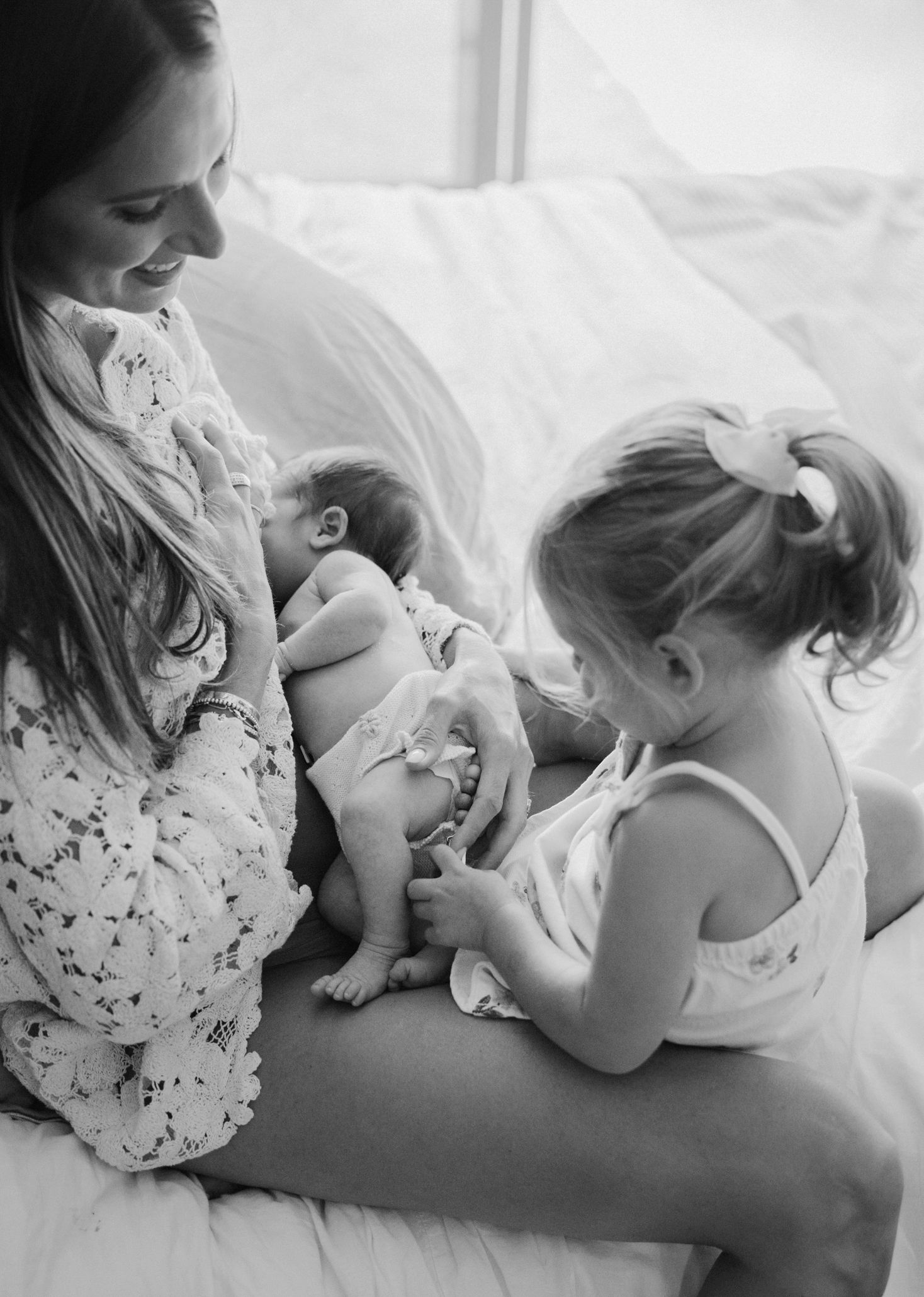 Black and white photo of a smiling mother holding a newborn while a toddler girl with a bow looks on tenderly.