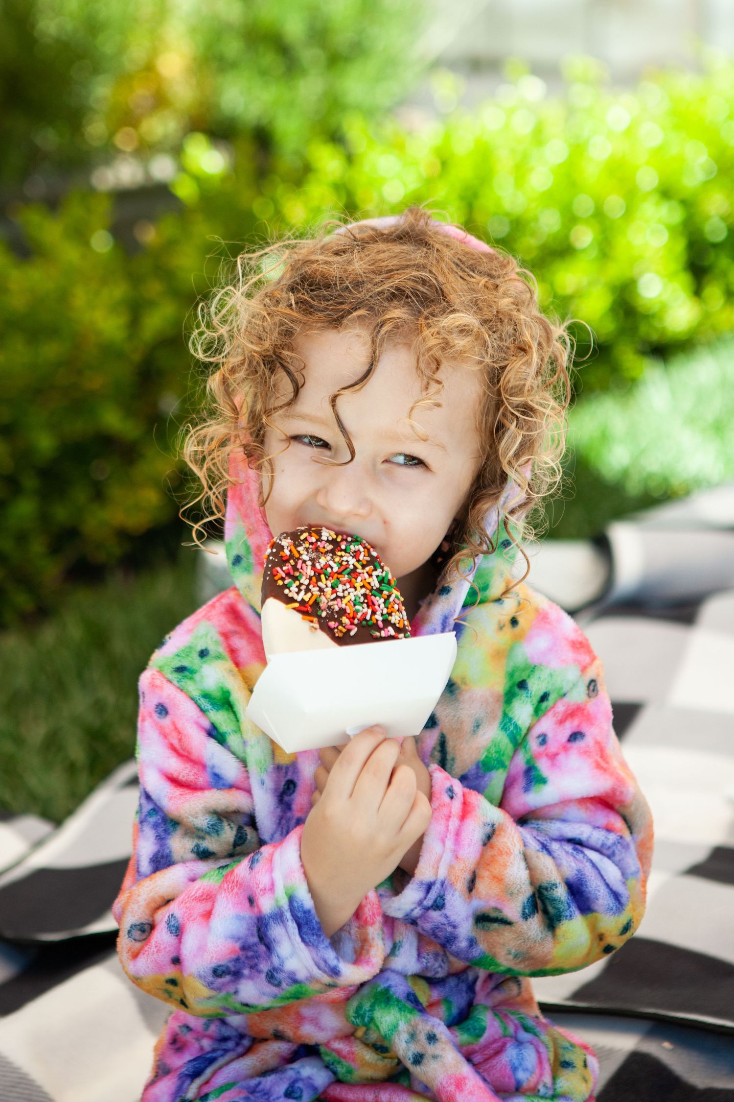 Curly-haired child in colorful robe eating a chocolate-dipped ice cream cone with rainbow sprinkles outdoors.