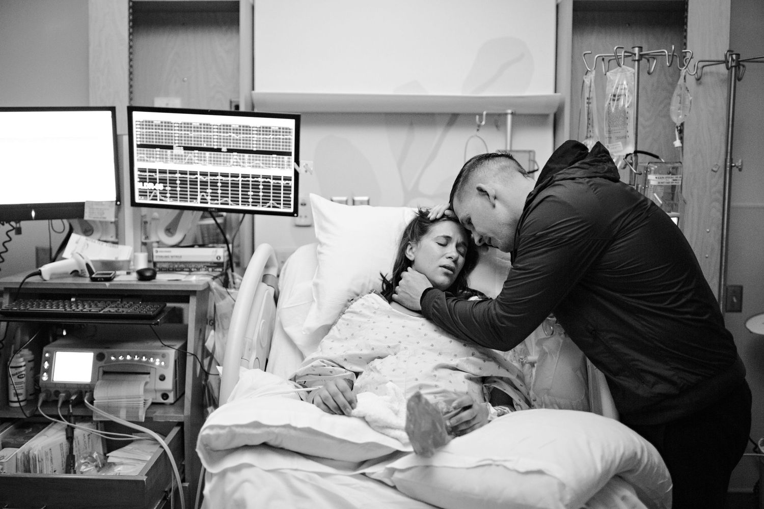 Black and white hospital photo of a man tenderly kissing a woman resting in a hospital bed with medical monitors nearby.