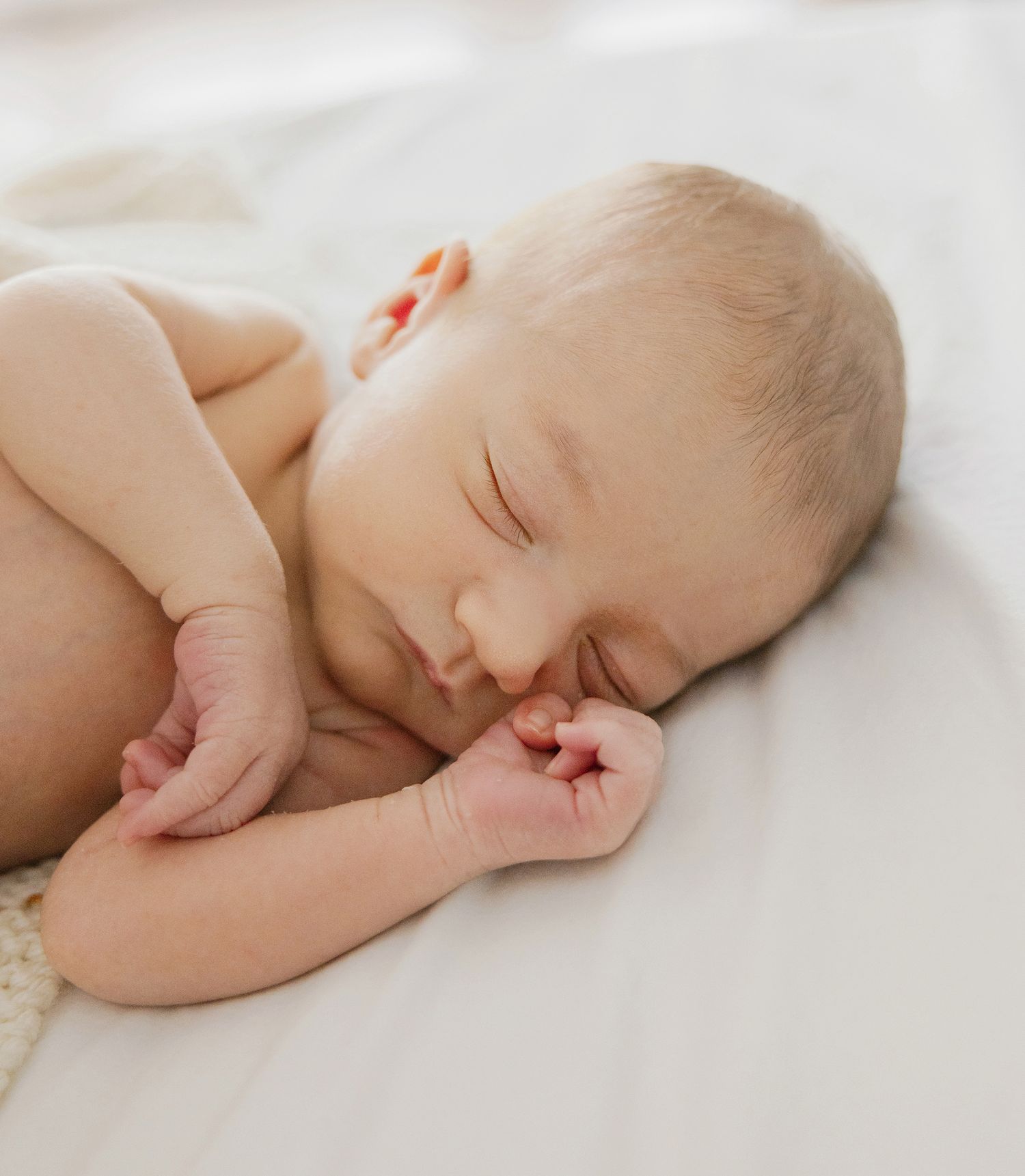 Peaceful newborn baby sleeping on white blanket with tiny fists curled, soft natural light highlighting delicate features.