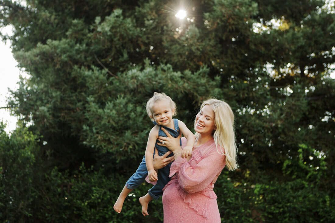 A smiling blonde woman in a pink dress holds a laughing toddler outdoors near lush green trees in warm sunlight.