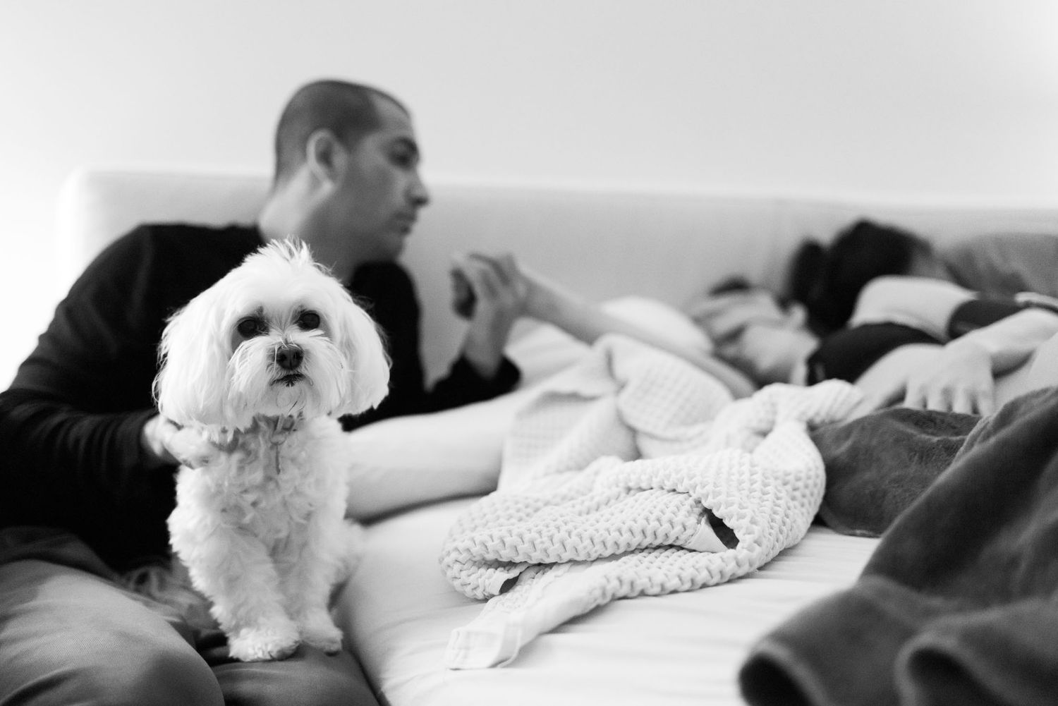 Black and white photo of a fluffy white dog sitting on a bed beside a new father and resting mother.