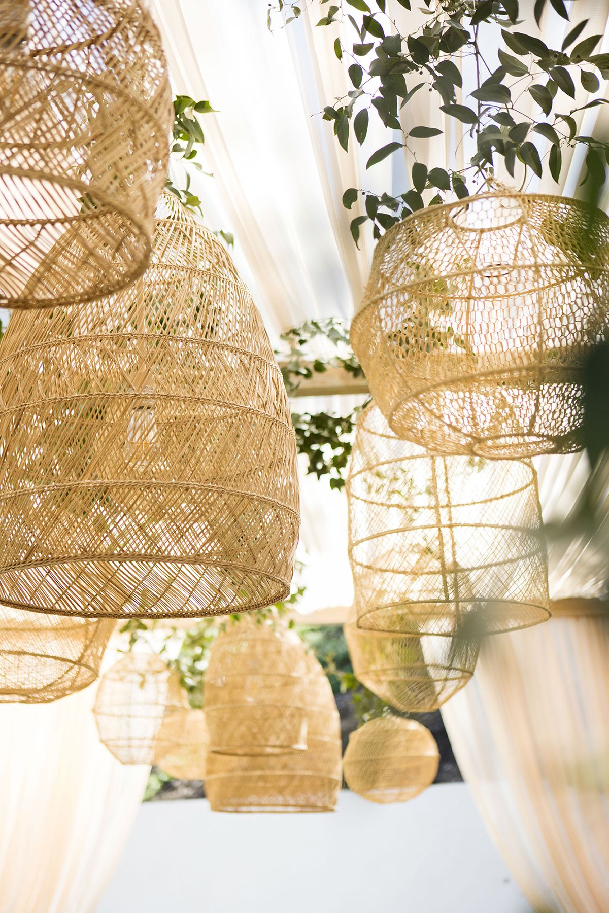 Woven rattan pendant light shades hanging from a white draped ceiling with green foliage at an outdoor event.