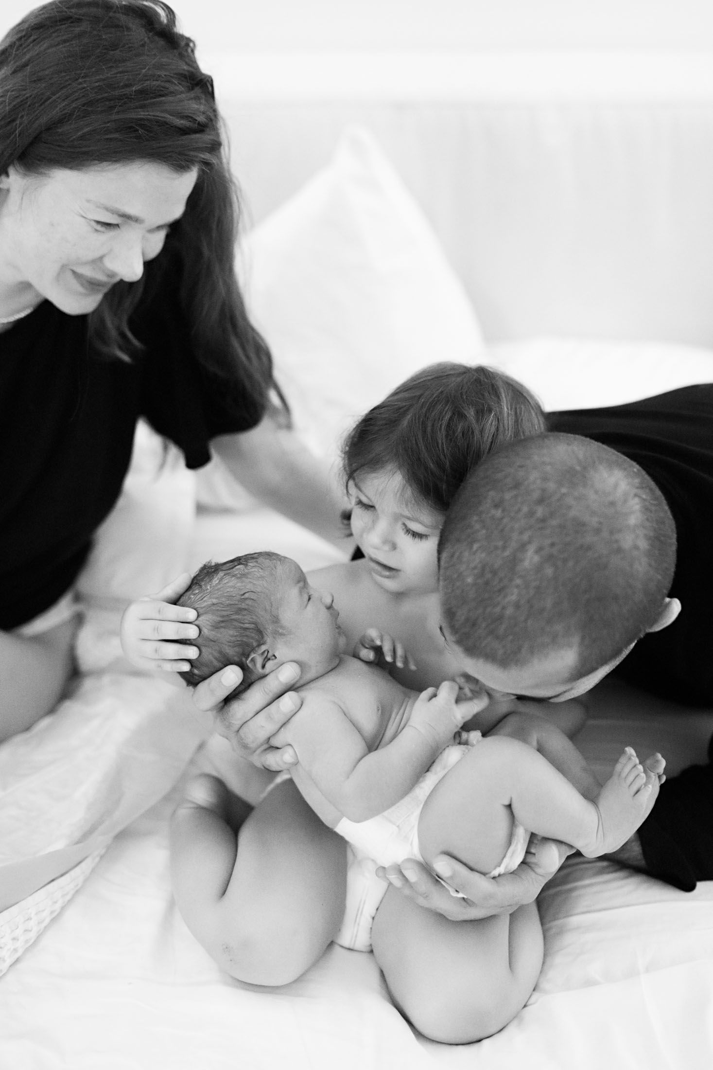 A mother and father smile as their toddler kisses a newborn baby in a heartwarming black and white photo.