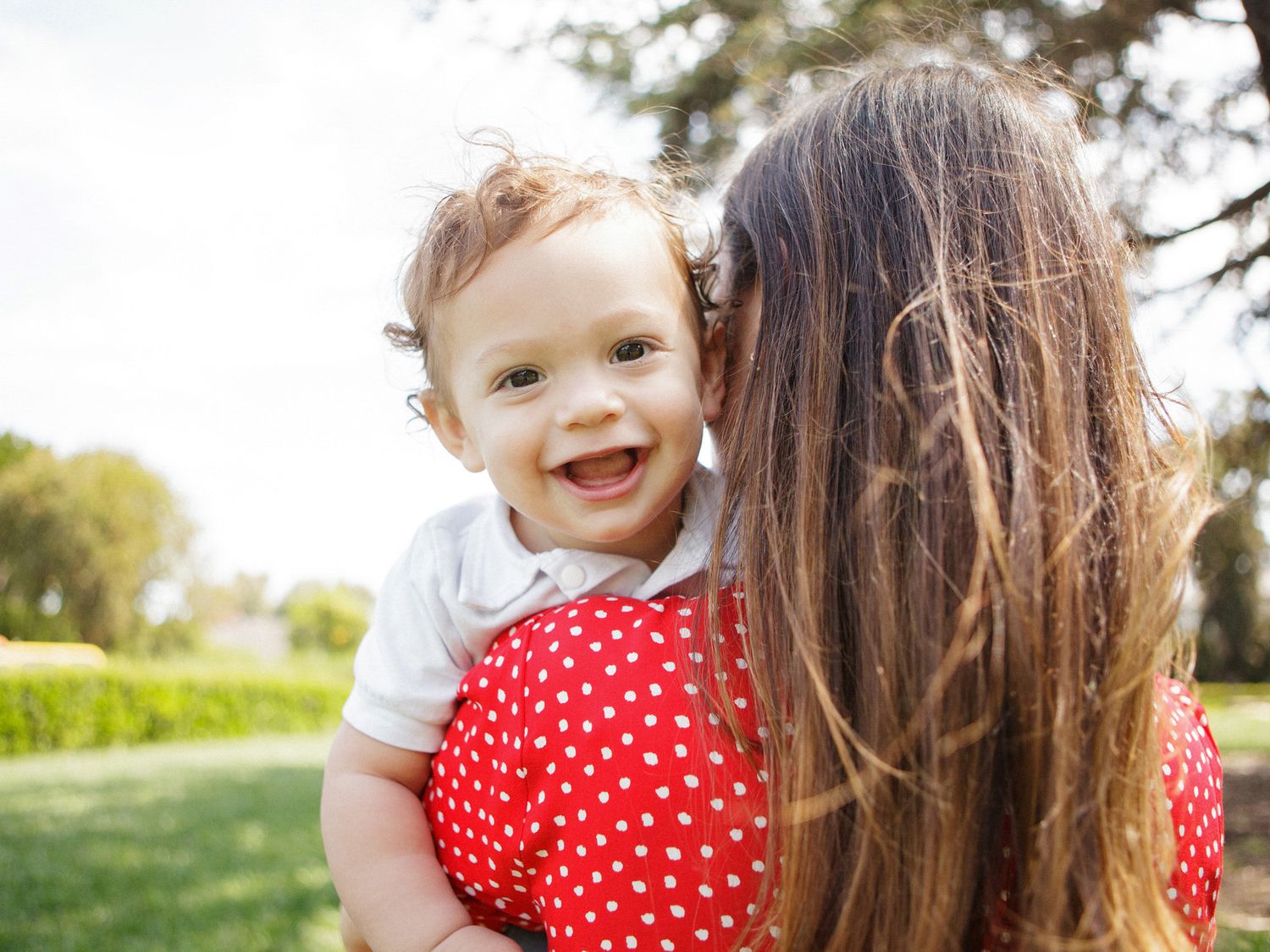 A happy baby smiles while being held by a woman in a red polka dot dress outdoors in a sunny park.