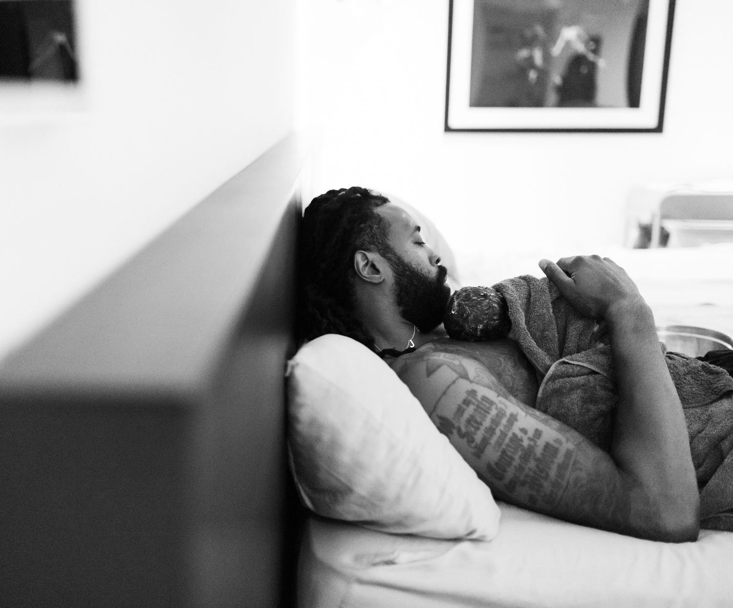 Black and white photo of a bearded man with tattoos resting on a bed with white pillows.