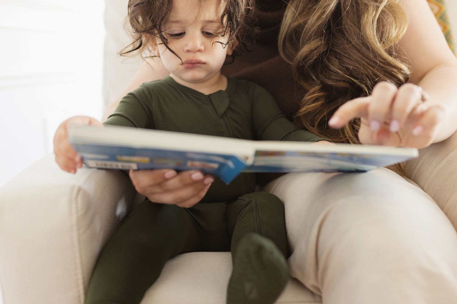 A toddler in green pajamas sits with a woman reading a colorful picture book together in a cozy chair.