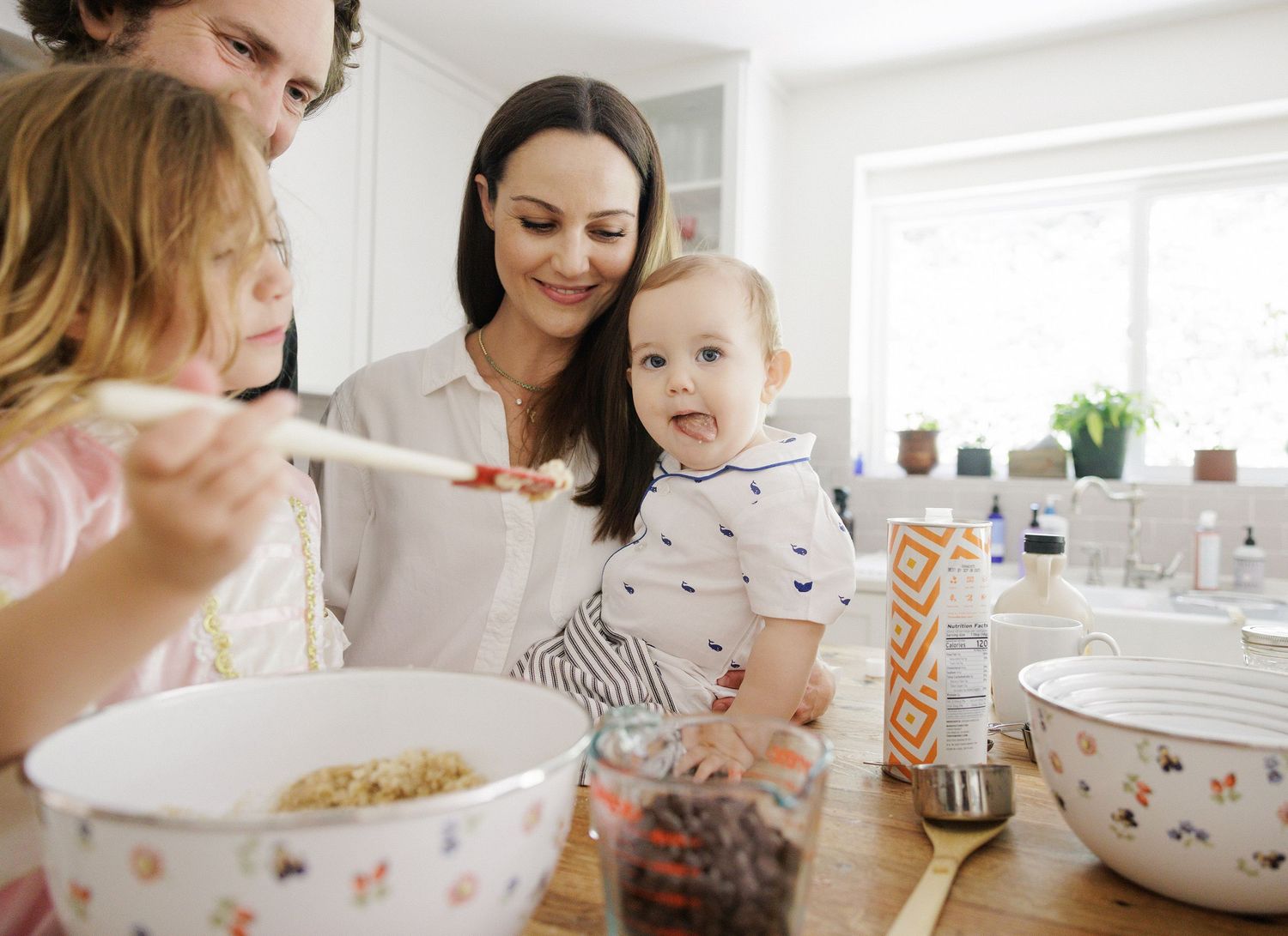 Family with baby and young girl baking together in a bright kitchen, mixing ingredients in a bowl.