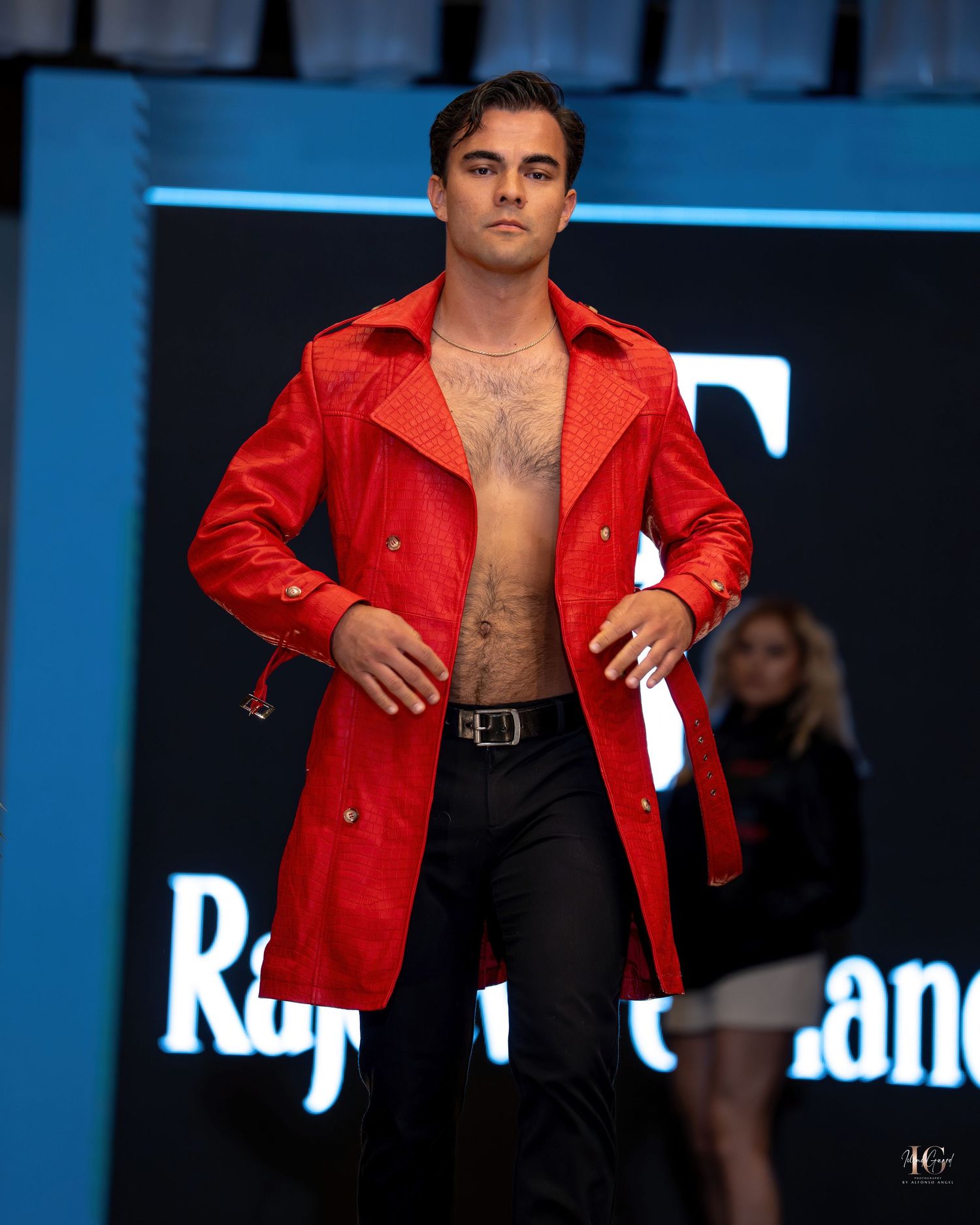 Model walks runway in bright red leather jacket with open front revealing bare chest against blue backdrop.